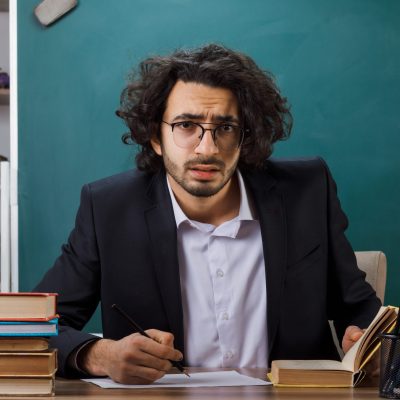 concerned male teacher wearing glasses writes something on paper sitting at table with school tools in classroom