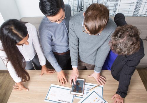 Closeup of four business colleagues discussing diagrams on tablet and papers. They are standing at table. Overhead view.